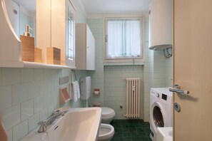Un bagno compatto con lavandino, WC e lavatrice, caratterizzato da rivestimenti piastrellati e una finestra.