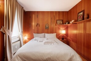 Un letto rifatto con lenzuola bianche, una lampada da comodino, una testiera in legno, un quadro incorniciato e uno scaffale con oggetti decorativi.
