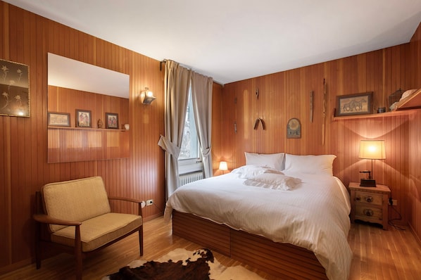 A bedroom with wooden paneling, a bed, a chair, a nightstand, a lamp, and a window with curtains.