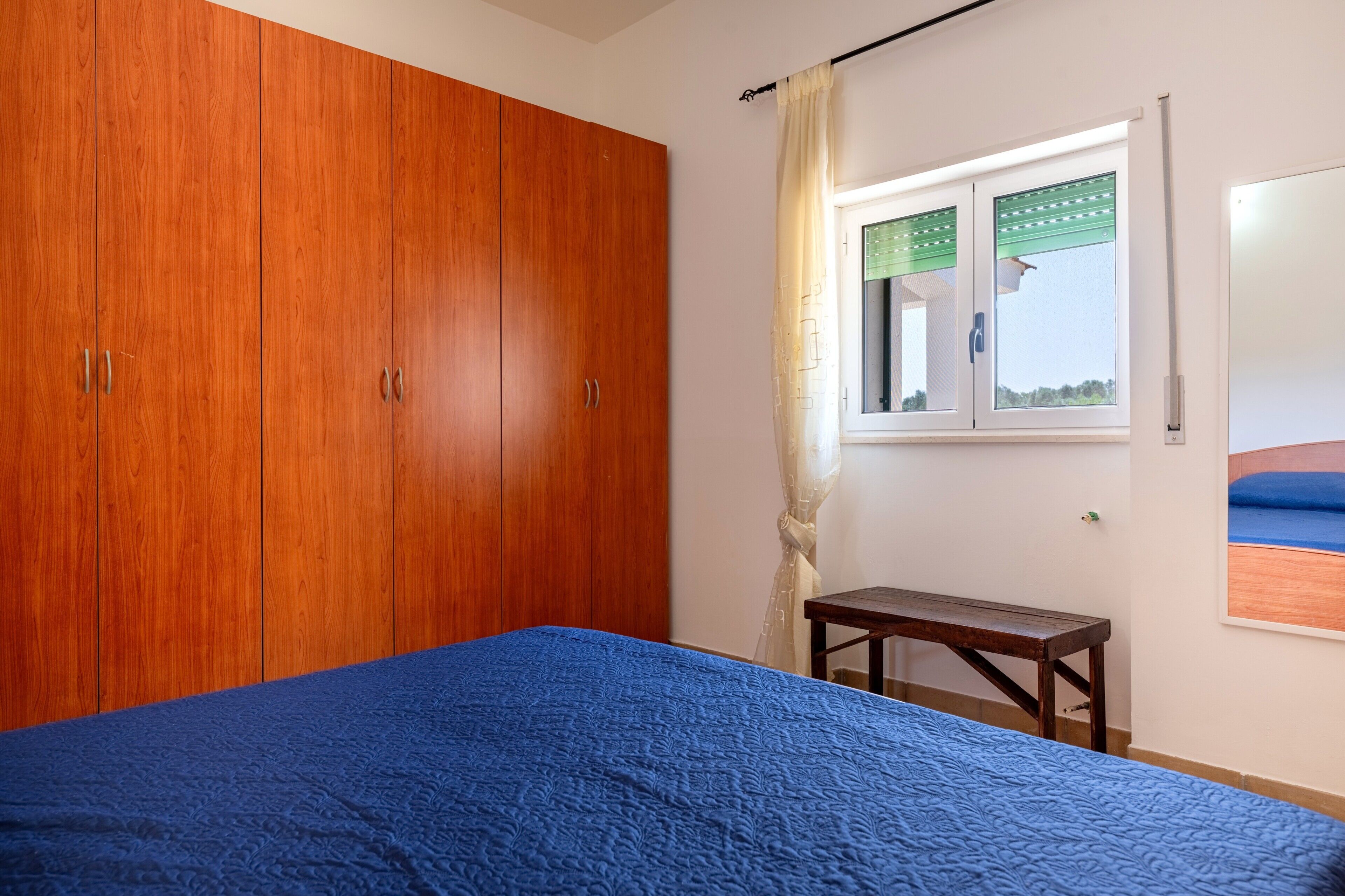 Un dormitorio con un armario grande, una cama con colcha azul, una mesita y un ventanal con vistas.