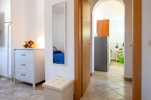 Een slaapkamer met een dressoir, een spiegel, een nachtkastje, een bed met een blauwe spreidek en uitzicht op de keuken.