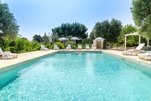 Piscina com espreguiçadeiras e guarda-sóis, cercada por árvores e vegetação.