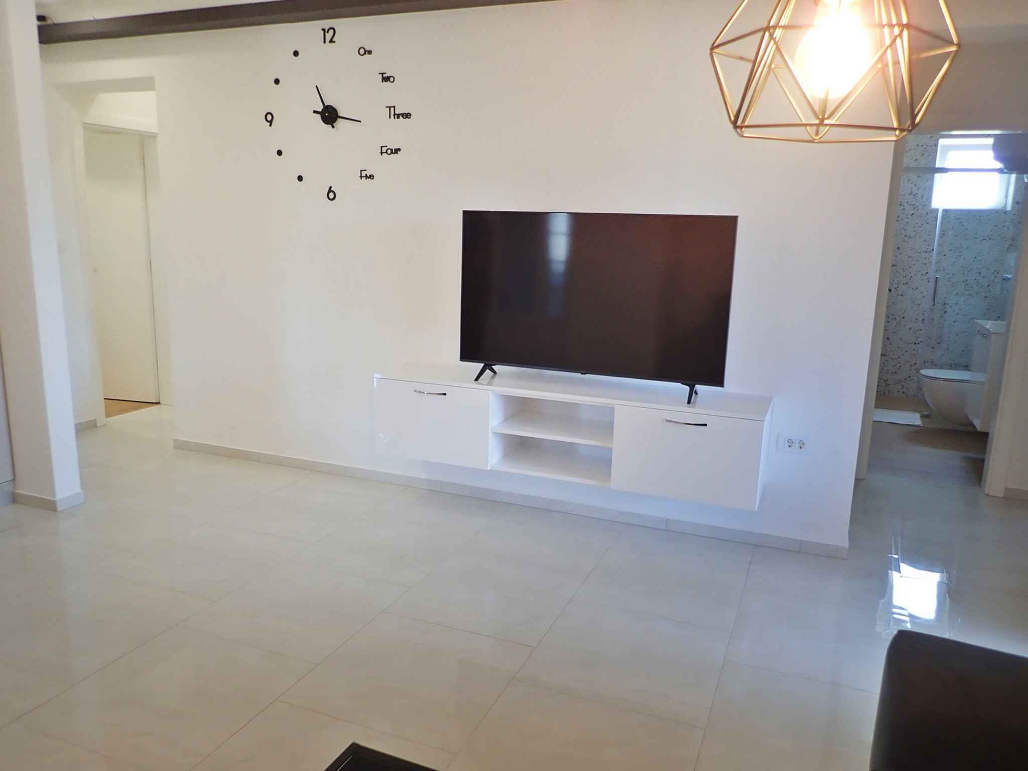 Ein modernes Wohnzimmer mit einem Flachbildfernseher an einer weißen Unterhaltungsecke, einer Wanduhr und einer geometrischen Pendelleuchte.