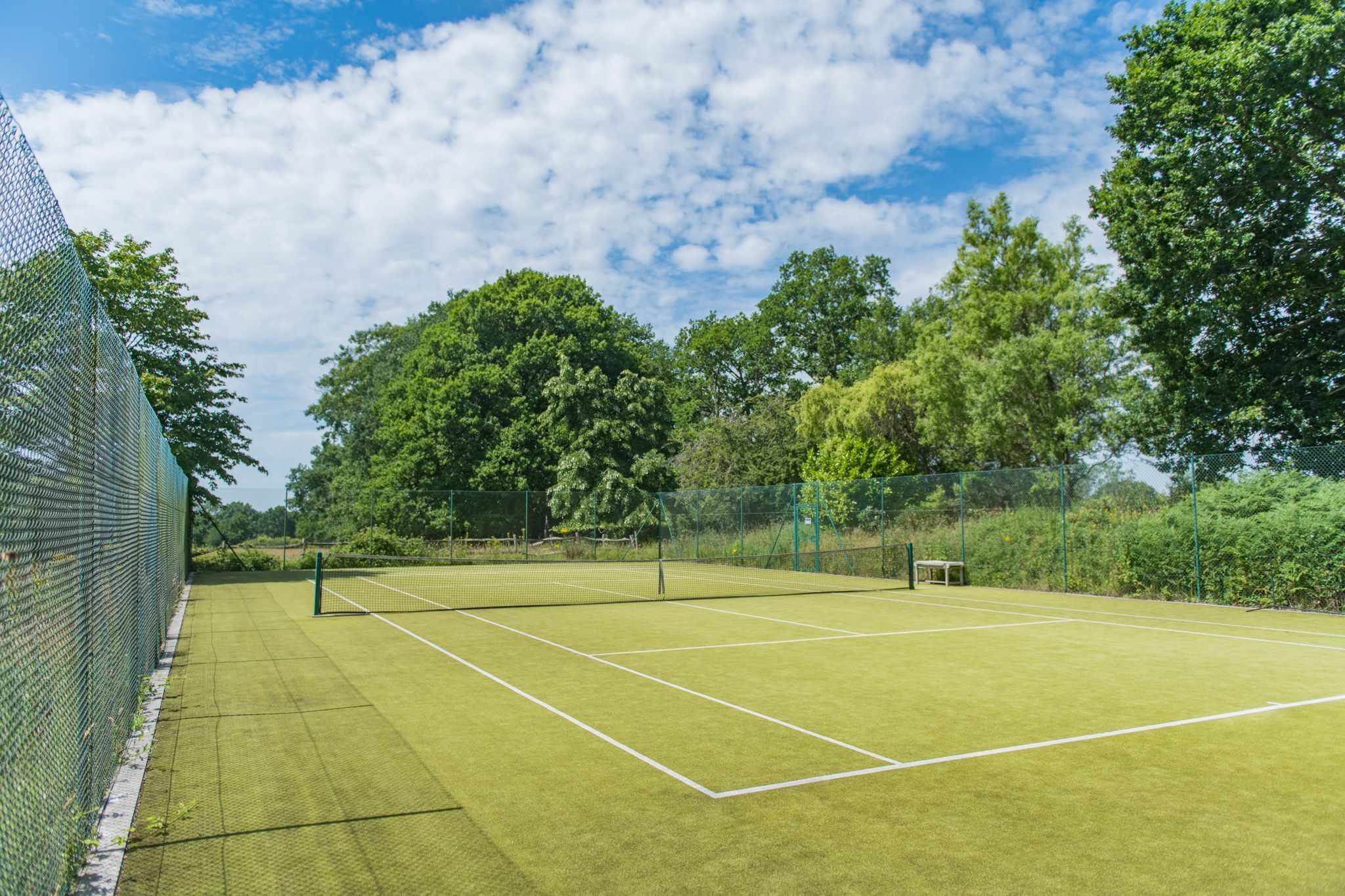 A tennis court with a net, surrounded by a green fence and trees.
