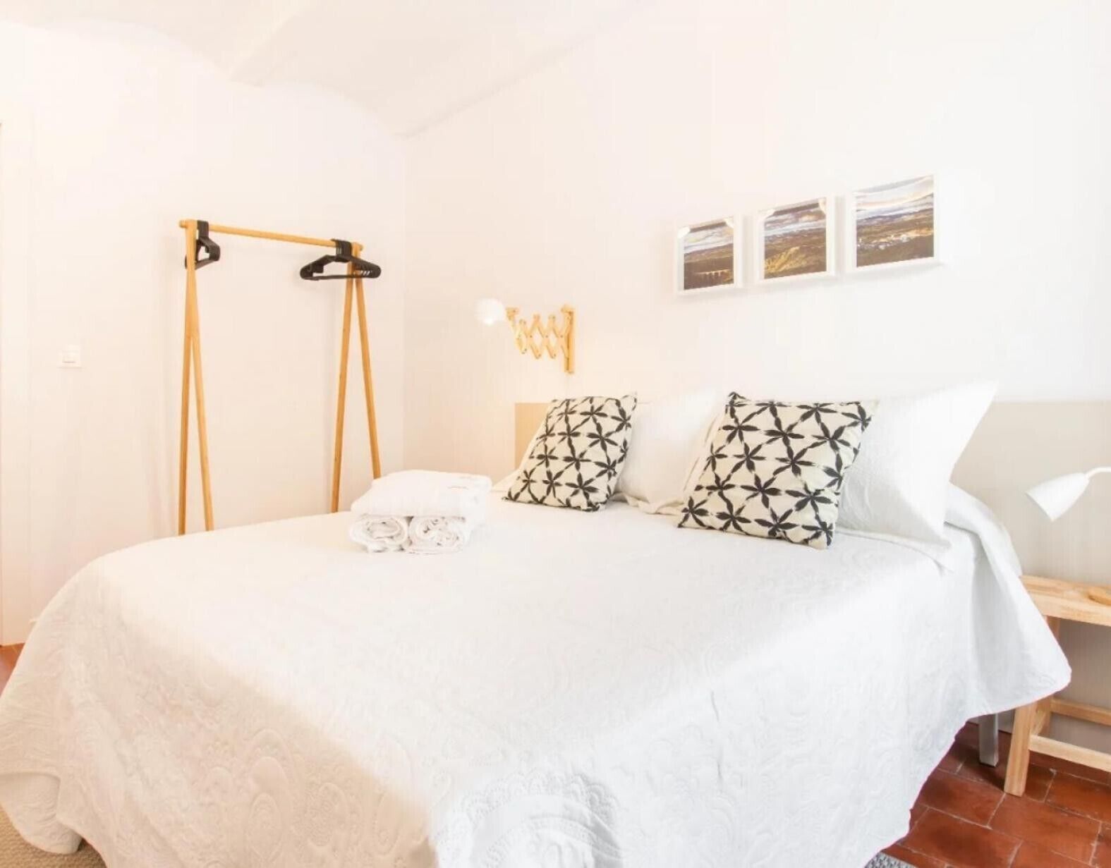 Una cama bien hecha con una colcha blanca y dos almohadas estampadas, una mesita de noche de madera con una lámpara, y un perchero en la pared con ropa.
