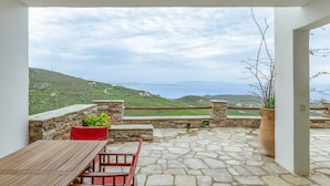 A stone-floored patio with a wooden table and chairs, a potted plant, and a view of green hills and the sea. - Holiday Home 'Modern New Tinos' with Sea View, Private Terrace and Wi-Fi (Tinos)