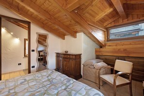 A bedroom with a wooden ceiling, a bed with a patterned bedspread, a wooden dresser, a chair, and a window with blinds.