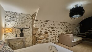 Una camera da letto con parete in pietra, un letto con cuscino decorativo, un comodino con lampada e una scala che conduce a un piano superiore.