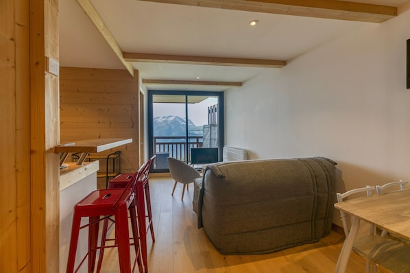 A room with a wooden ceiling, a sofa, a dining table, and a view of mountains through a large window.