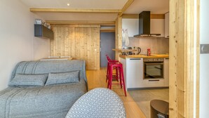 A compact room with a sofa, a dining area with a red chair, a kitchen with a stove, and a wooden ceiling. - Apartment 'Alpe D'huez' with Mountain View and Balcony (Huez)