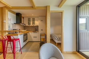Una cocina compacta con vigas de madera, una zona de comedor con sillas rojas, un sillón estampado y una cama en la habitación contigua.