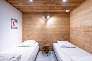 Um quarto pequeno com duas camas, paredes de madeira, um banquinho e um cartaz na parede.
