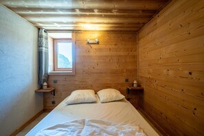 A wooden cabin bedroom with a bed, pillows, a nightstand, a window, and a small shelf. - Apartment 'La Bergerie' with Mountain View, Private Terrace and Wi-Fi (La Giettaz)