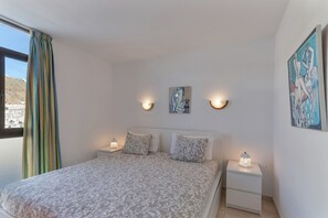 Een slaapkamer met een bed, twee nachtkastjes met lampen, een raam met gordijnen, en twee aan de muur bevestigde kunstwerken.