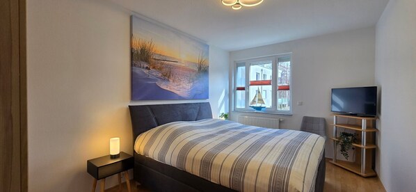 A bedroom with a bed, a painting, a TV, and a window with a view of buildings.