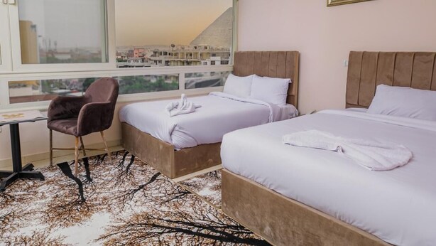A hotel room with two beds, a desk, a chair, and a large window with a city view. - Mak Pyramids View (Giza)