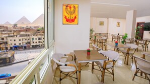 A dining area with tables and chairs, a view of the pyramids, and a wall with a colorful artwork. - Mak Pyramids View (Giza)
