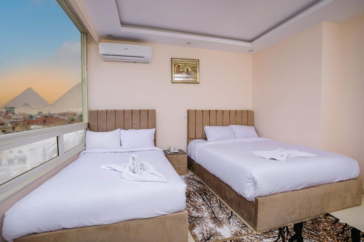 A hotel room with two beds, a view of the pyramids, an air conditioner, and a framed picture on the wall.