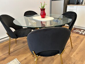 A round glass table with four black chairs with gold legs, a red vase with flowers, and a white cabinet in the background.
