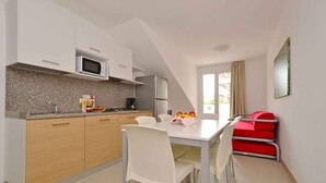 A modern kitchen with white cabinets, a microwave, and a refrigerator. There is a dining table with white chairs and a fruit bowl. - Holiday apartment with air conditioning, internet, balcony and pool (Lignano)