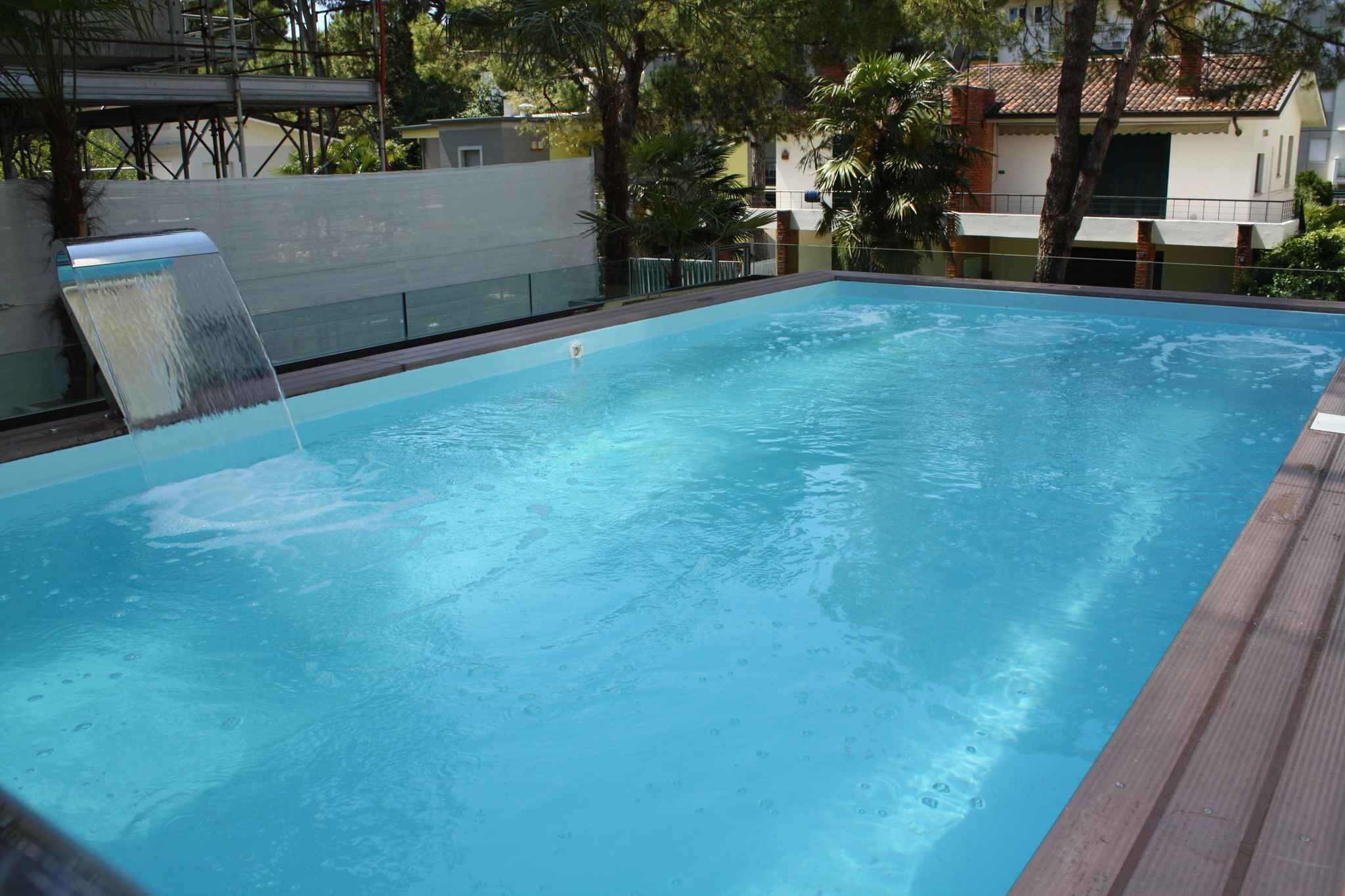Piscina rectangular con una cascada en un lateral, rodeada por una terraza de madera y árboles.