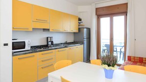 Una cocina compacta con armarios amarillos, una encimera blanca, microondas, estufa y refrigerador. Hay una mesa de comedor con sillas amarillas y una planta en maceta.