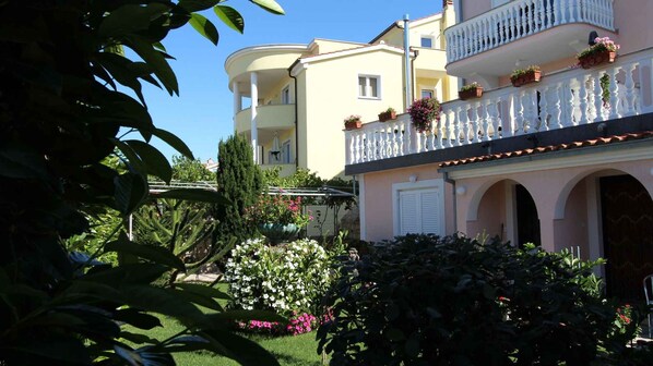 Un immeuble à plusieurs étages doté de balcons, un jardin avec différentes espèces de plantes et un ciel d’un bleu limpide.
