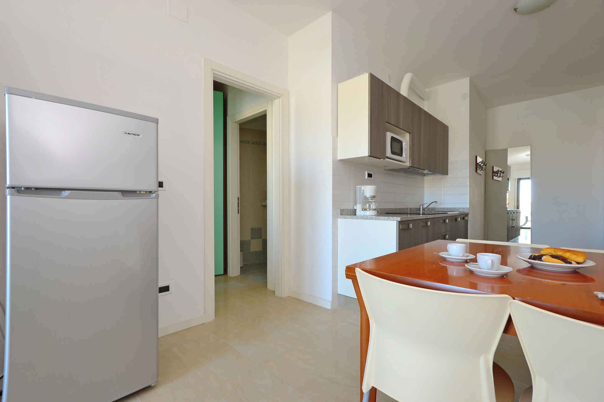 Cocina con refrigerador, microondas y cafetera. Hay una mesa de comedor con sillas y un plato de comida.