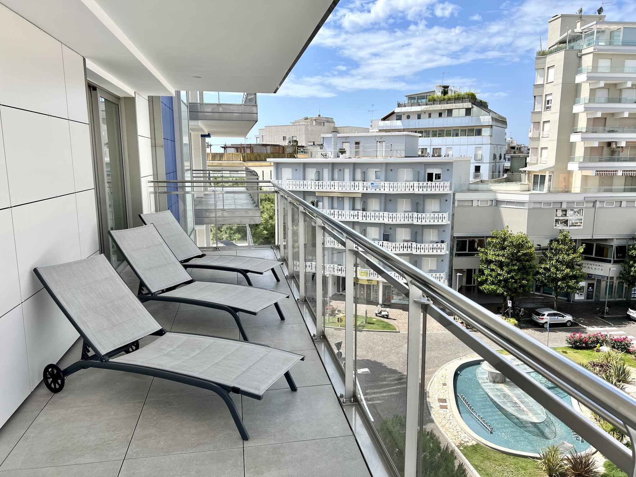A balcony with three lounge chairs, a pool, and a view of surrounding buildings.