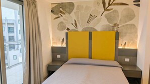 A bedroom with a bed, a large window with curtains, a wall with abstract art, and a view of buildings outside.