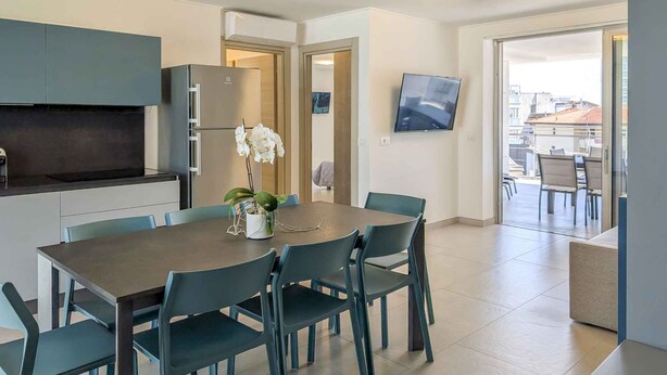 A modern kitchen with a built-in oven, a dining table with chairs, a potted orchid, and a flat-screen TV mounted on the wall.