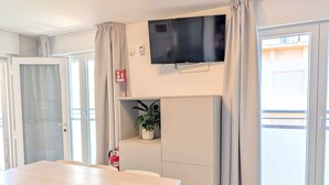 A modern room with a flat-screen TV, a dining table, chairs, a potted plant, and a fire alarm. - Holiday apartment with balcony and air conditioning (Lignano)