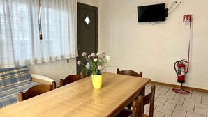 Un coin repas avec une table en bois, des chaises, un vase jaune rempli de fleurs, un téléviseur fixé au mur et un extincteur.