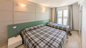 A hotel room with two beds, a green headboard, a nightstand, and a window with curtains. - Holiday flat with balcony and air conditioning (Lignano)