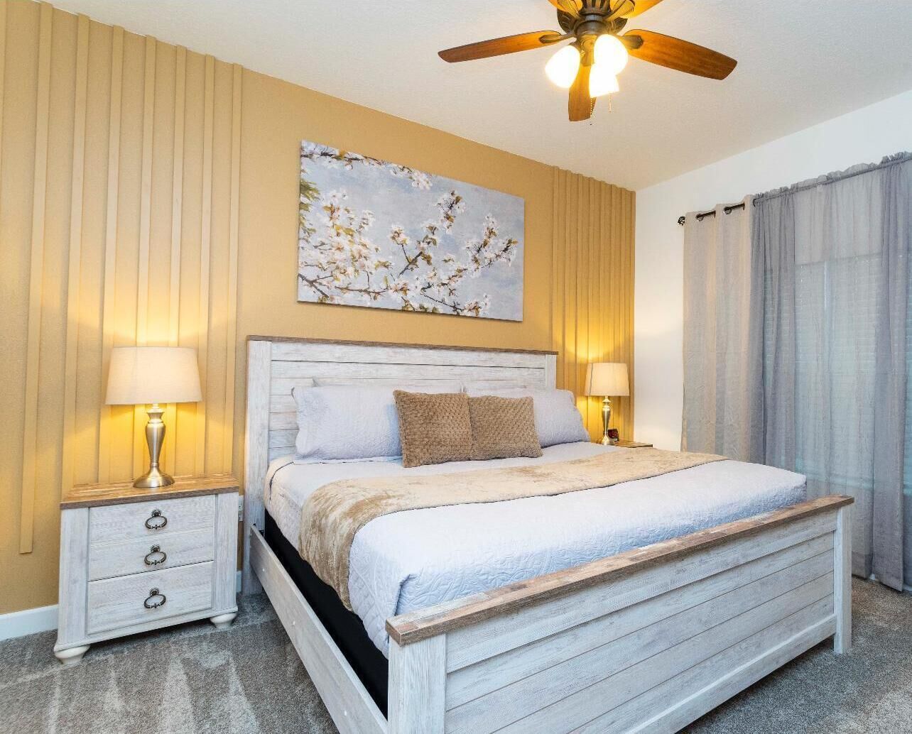 A bedroom with a bed, two bedside tables, a ceiling fan, a painting, and curtains.
