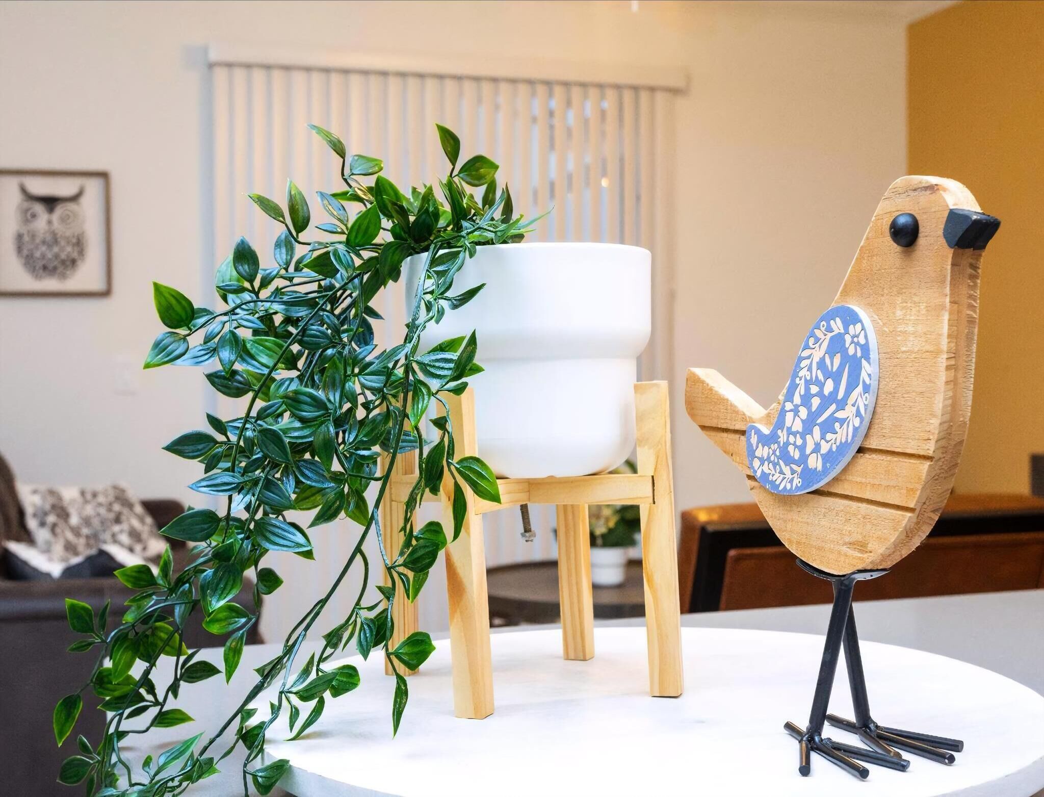 Um quarto com uma mesa branca, uma cadeira de madeira, uma planta em vaso e uma escultura de pássaro de madeira.