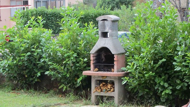 A stone outdoor pizza oven with a fire burning inside, surrounded by lush green bushes.