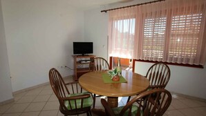 Un comedor con una mesa redonda de madera, cuatro sillas de madera, un televisor sobre un mueble bajo y una ventana con persianas.