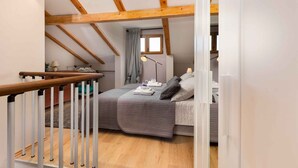 Camera da letto con soffitto a falde, travi in legno, un letto con copripiumino grigio, una lampada da comodino e una finestra con tenda.