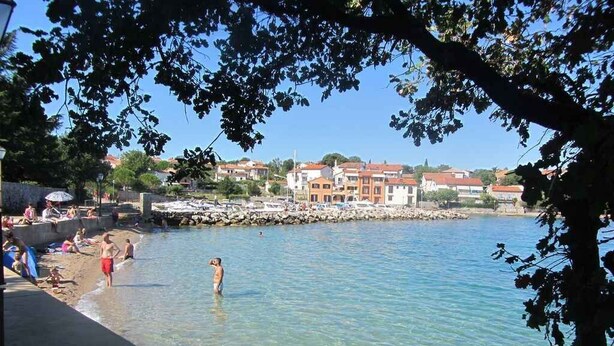 Una battigia limpida con persone che nuotano e passeggiano lungo la riva, un molo frastagliato e edifici sullo sfondo.