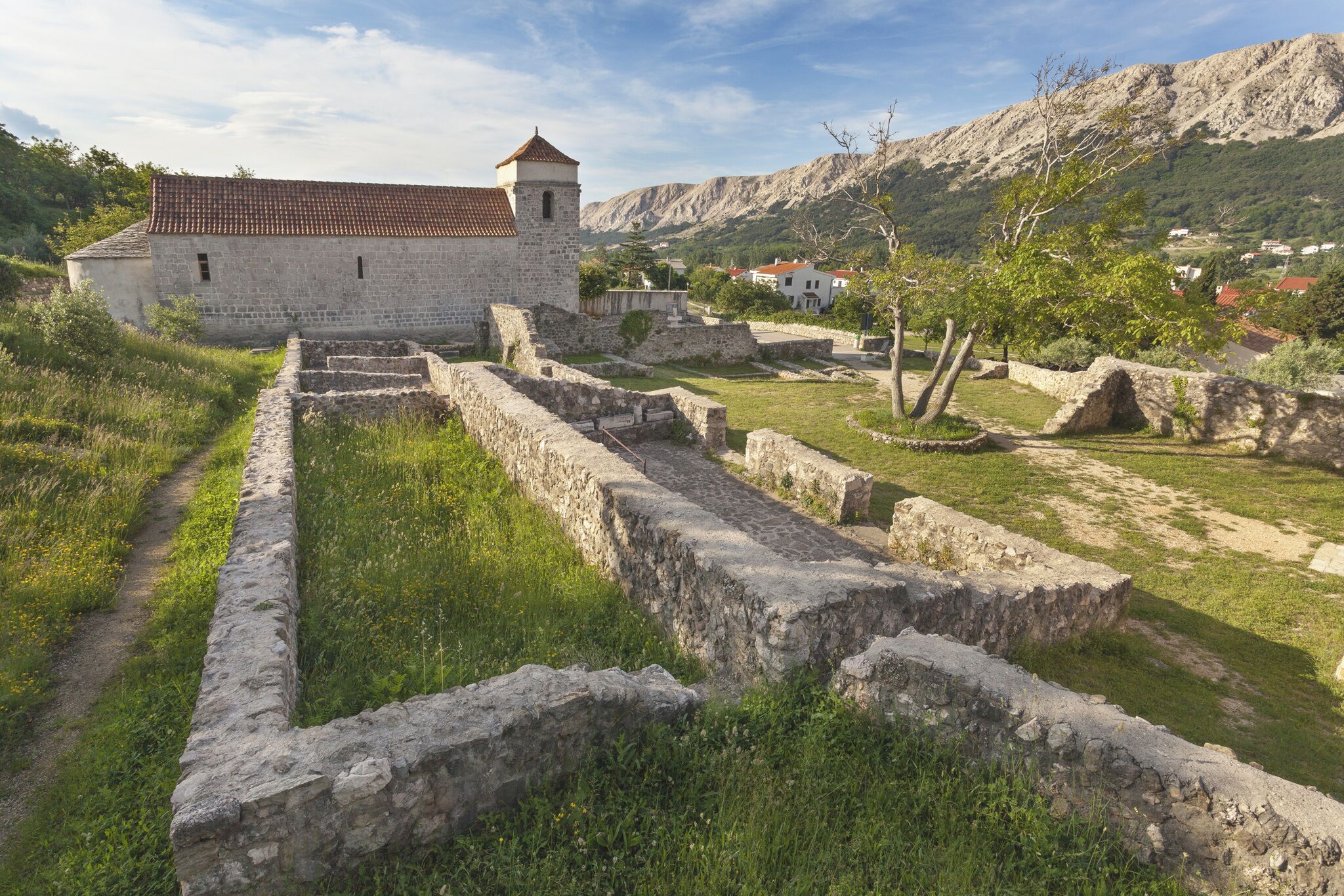 Un edificio de piedra con techo de tejas rojas, rodeado de ruinas y un paisaje montañoso.