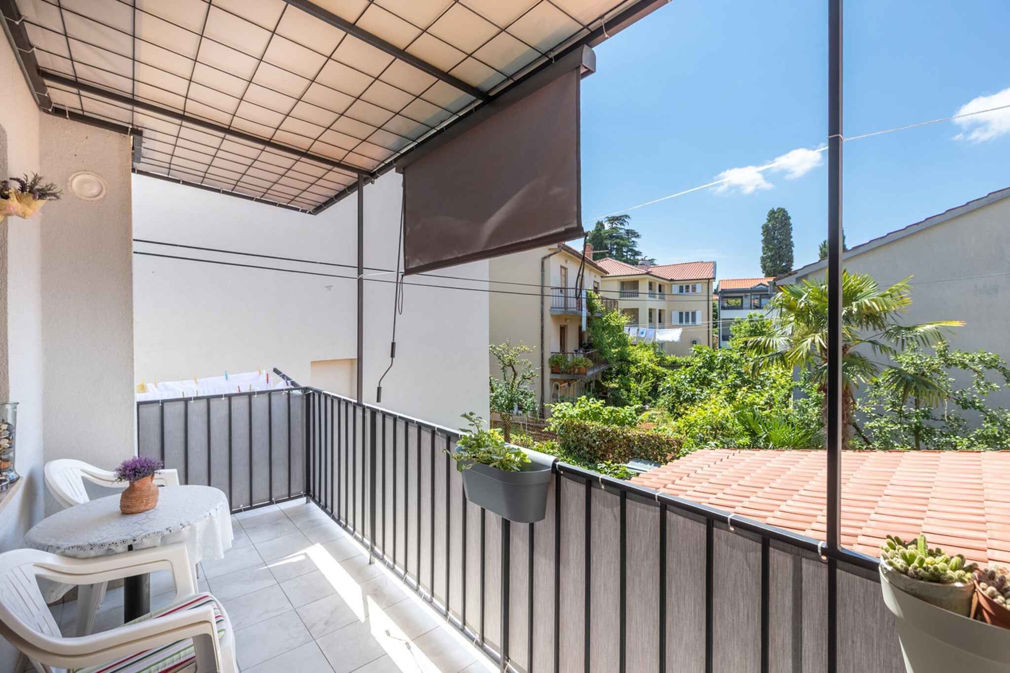 Un balcon avec une table et des chaises, une plante suspendue, et une vue sur les bâtiments et la verdure environnants.