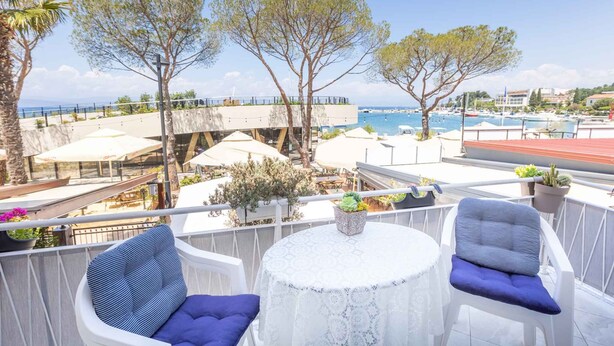 Ein Balkon mit weißen Stühlen, einem runden Tisch und Blick auf das Meer und Gebäude.