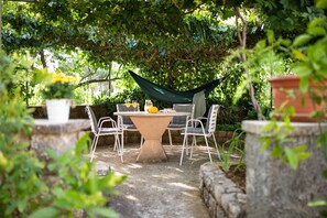 A garden patio with a table and chairs, surrounded by greenery and a hammock.