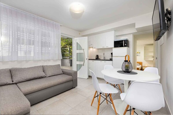 A modern living room with a grey sofa, a dining area with white chairs, a kitchen with white cabinets, and a TV mounted on the wall.