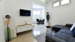 A modern living room with a grey sofa, a flat-screen TV mounted on the wall, a dining area with a table and chairs, and a small side table with a plant.