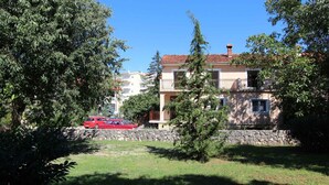 Edifício residencial com parede de pedra, um carro vermelho estacionado na frente e vegetação exuberante.