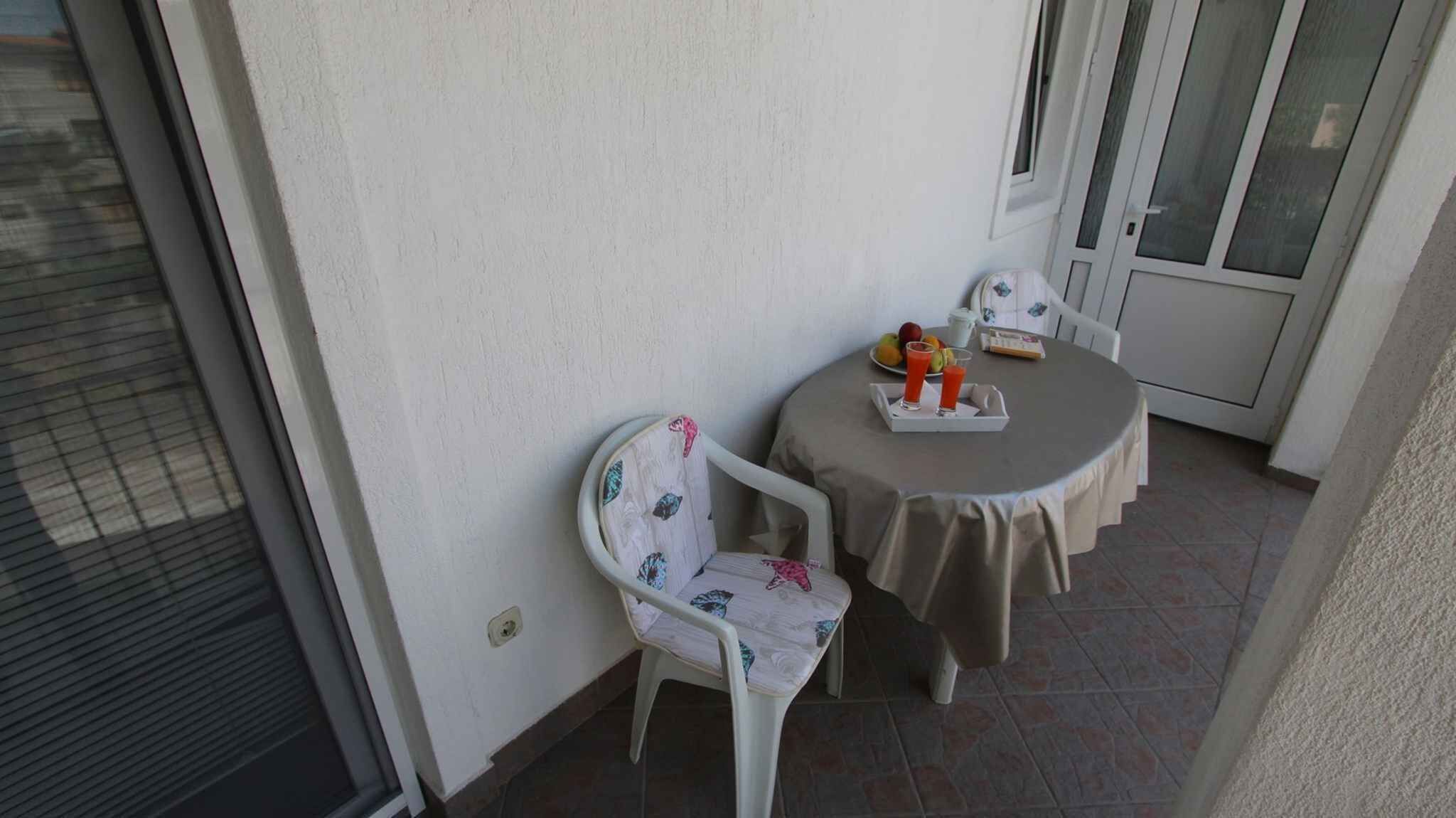 Un pequeño balcón con una silla blanca, una mesa, una puerta y suelo de baldosas.