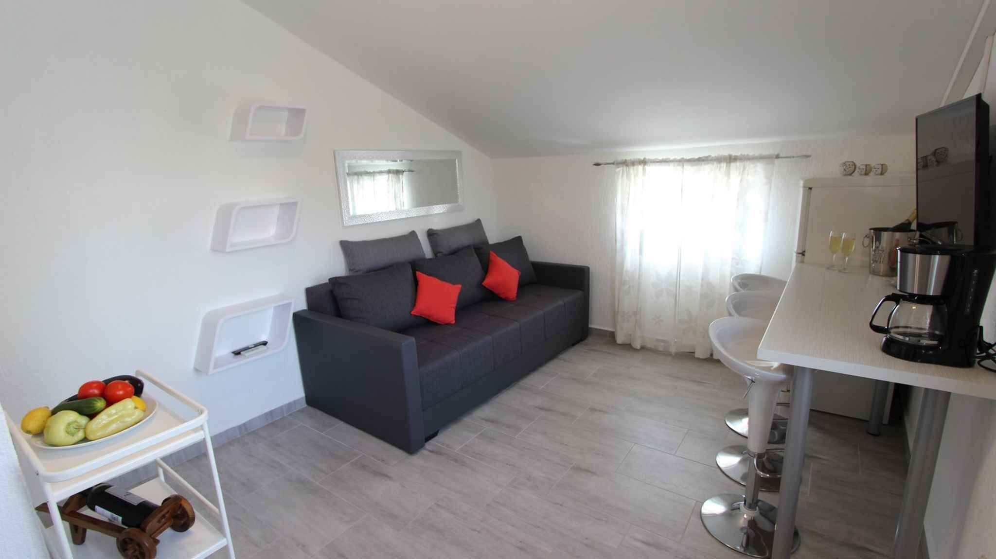 A modern living room with a dark sofa, red cushions, a white coffee table with fruits, a kitchen area with a coffee maker, and a bathroom with a mirror and toilet.