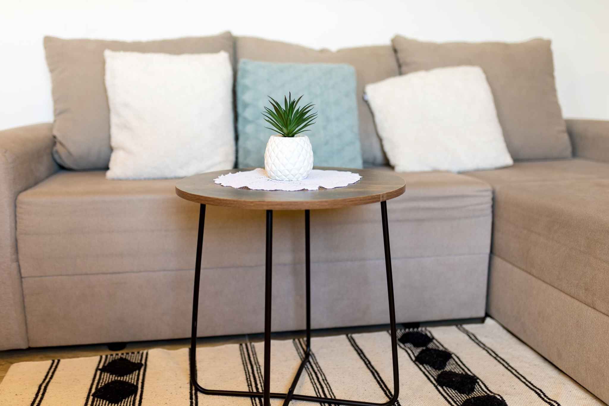 Een kleine ronde tafel met een wit, geribbeld blad en zwarte metalen poten, geplaatst op een tapijt met een patroon, voor een beige bank met drie kussens.